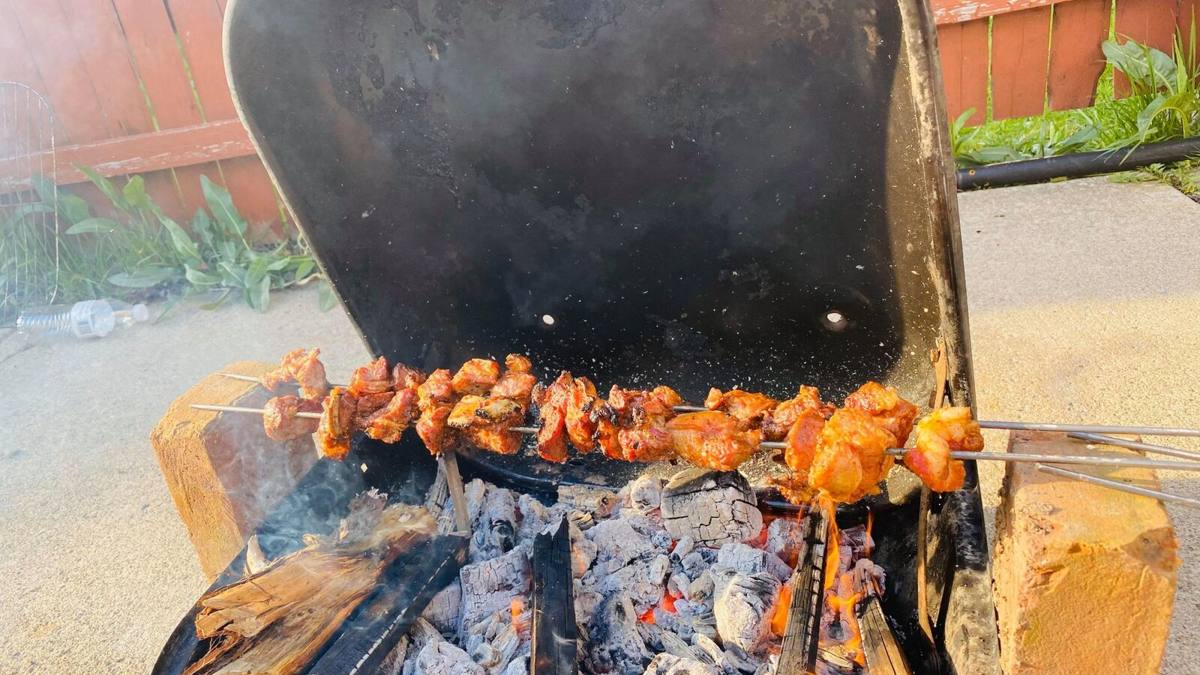 Skewers of marinated chicken laid across a wood fire in a back garden, the grill lid propped open behind them.
