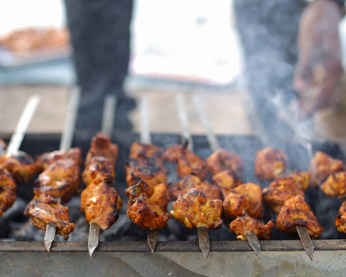 A long row of chicken skewers turning on a grill, with people standing just behind it in the smoke.