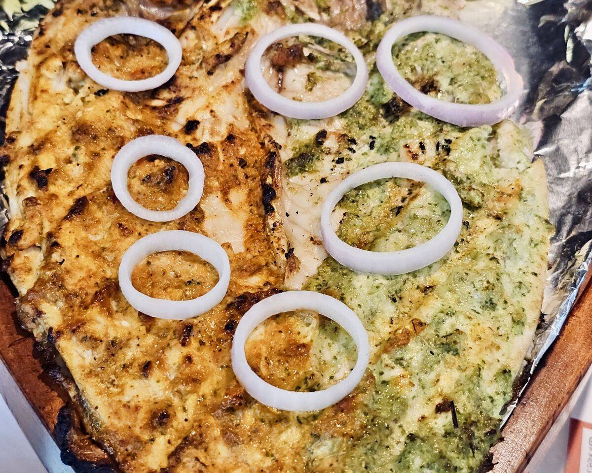 A whole fish grilled under a green marinade, laid out on a tray with rings of raw onion.