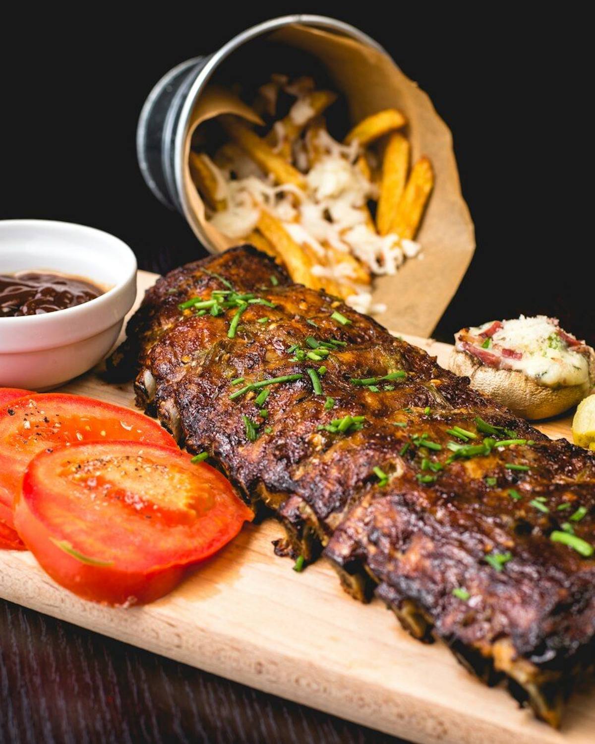A rack of smoked ribs on a serving board with pickles and sliced tomato.