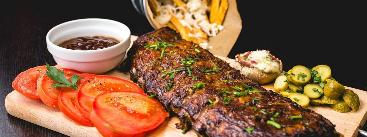 A rack of smoked ribs on a serving board with pickles and sliced tomato.