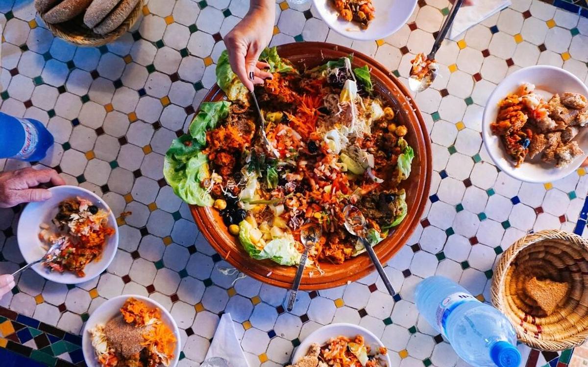 Hands reaching in over a tiled table crowded with shared bowls, plates and drinks.